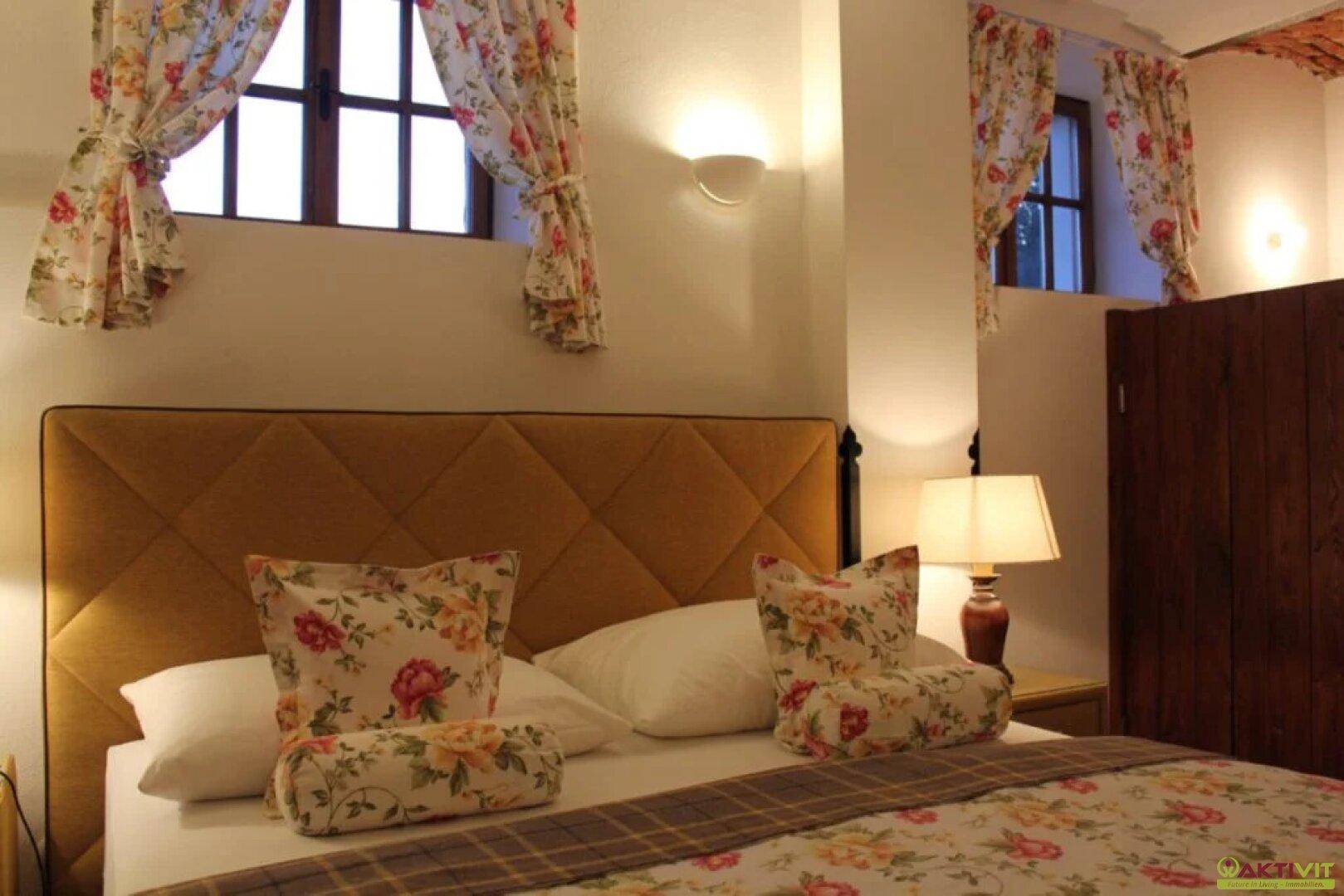Gemütliches Schlafzimmer mit floralen Vorhängen, gepolstertem Kopfteil und warmem Licht.