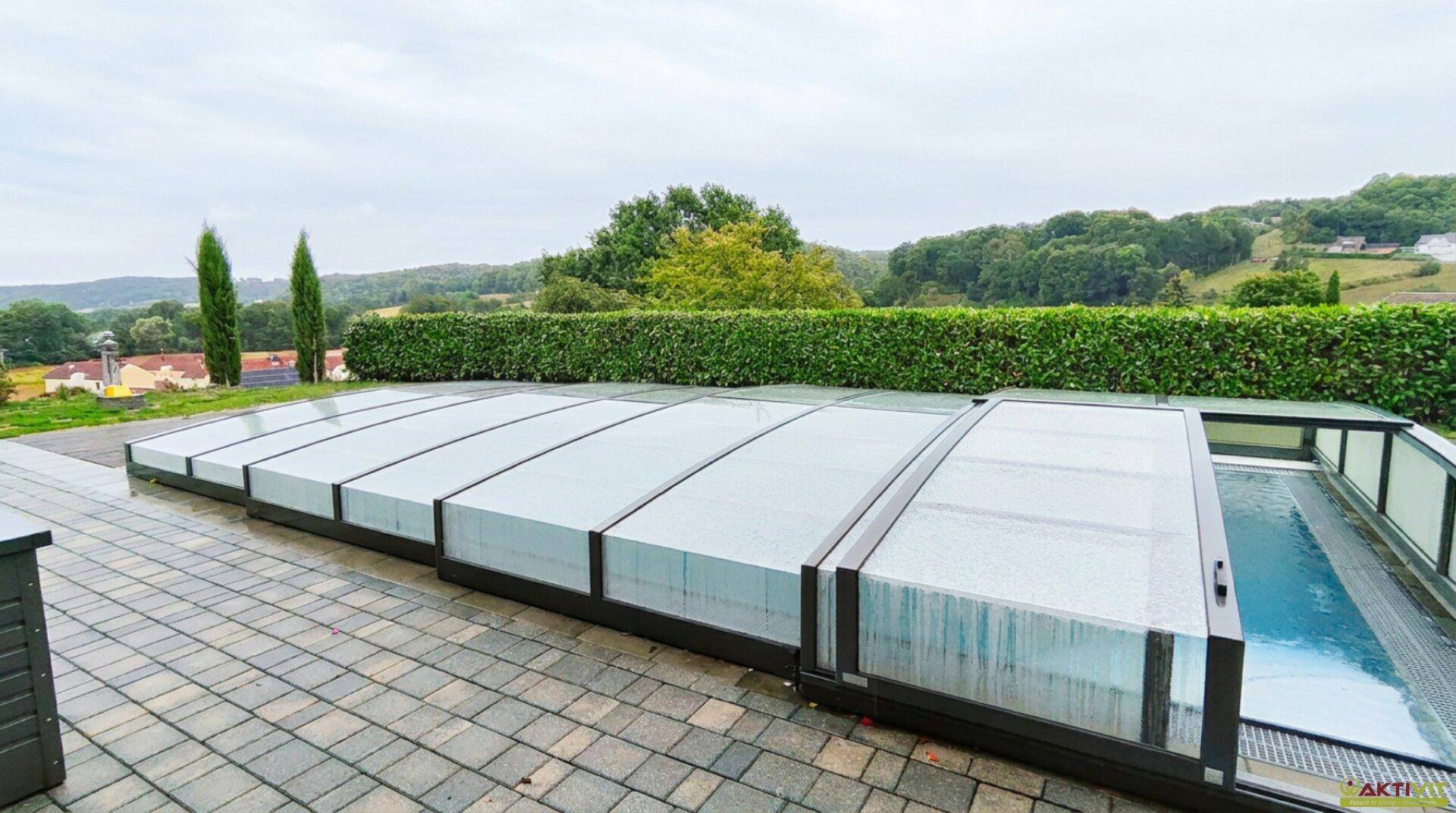 Überdachter Swimmingpool-Bereich mit gepflasterter Terrasse und Blick auf die grüne Landschaft.