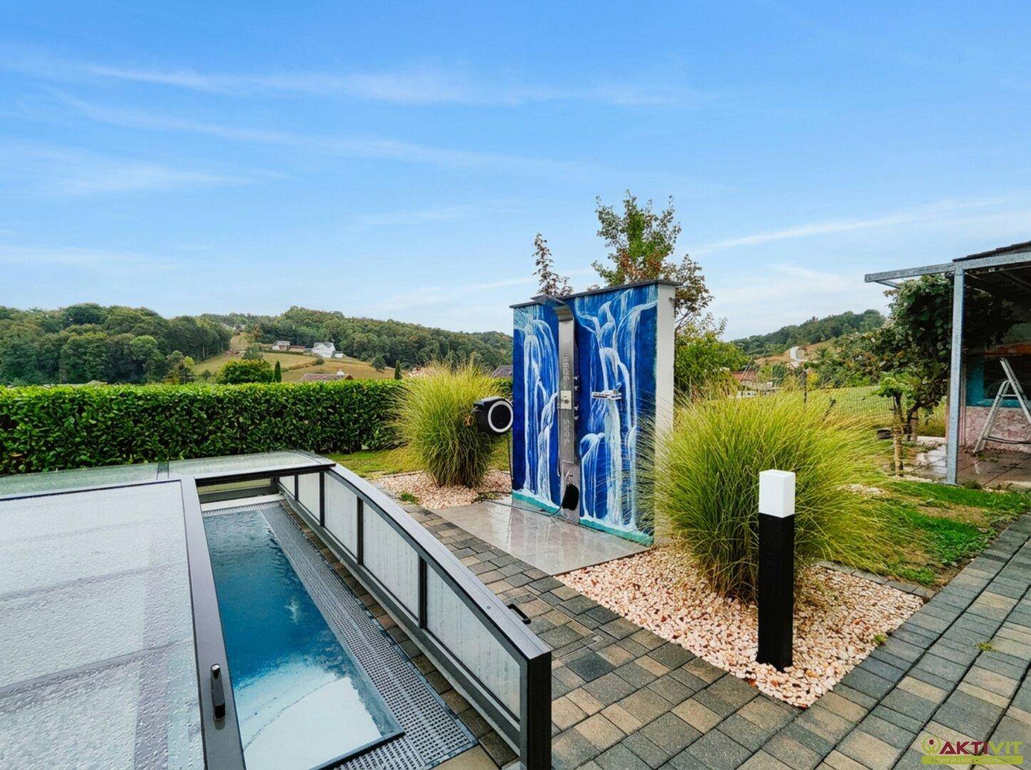Außenbereich mit überdachtem Pool, dekorativer Dusche und Kieswegen, umgeben von Grün.