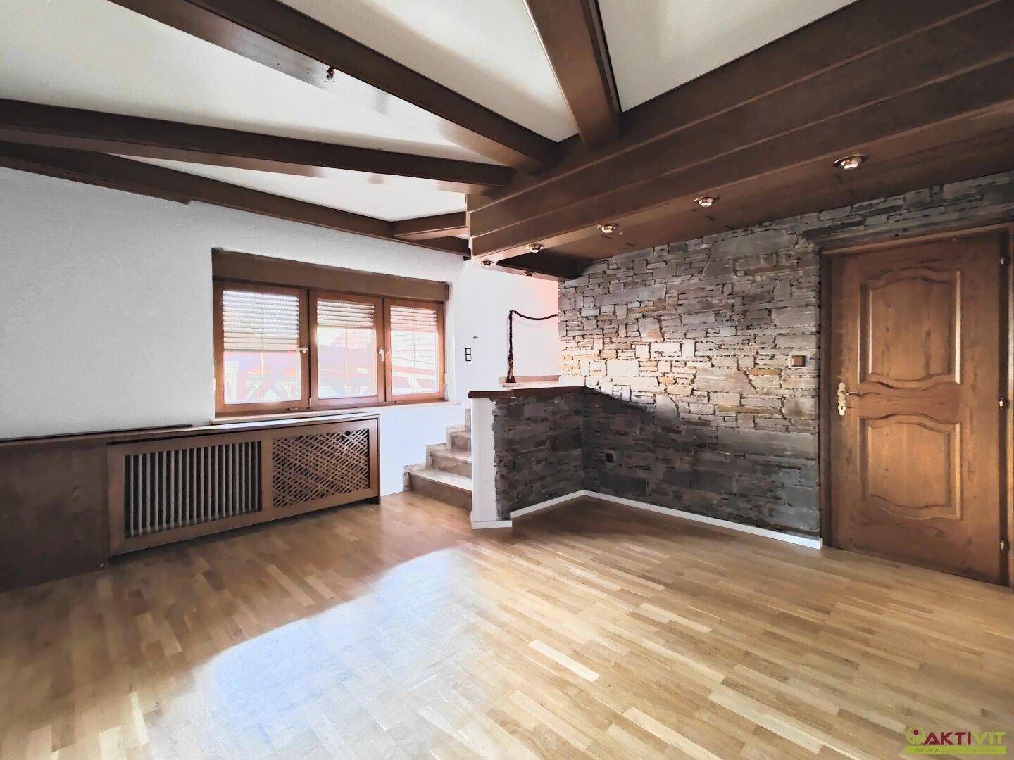 Geräumiges Wohnzimmer mit Holzbalkendecke, Parkettboden und einer dekorativen Steinwand.