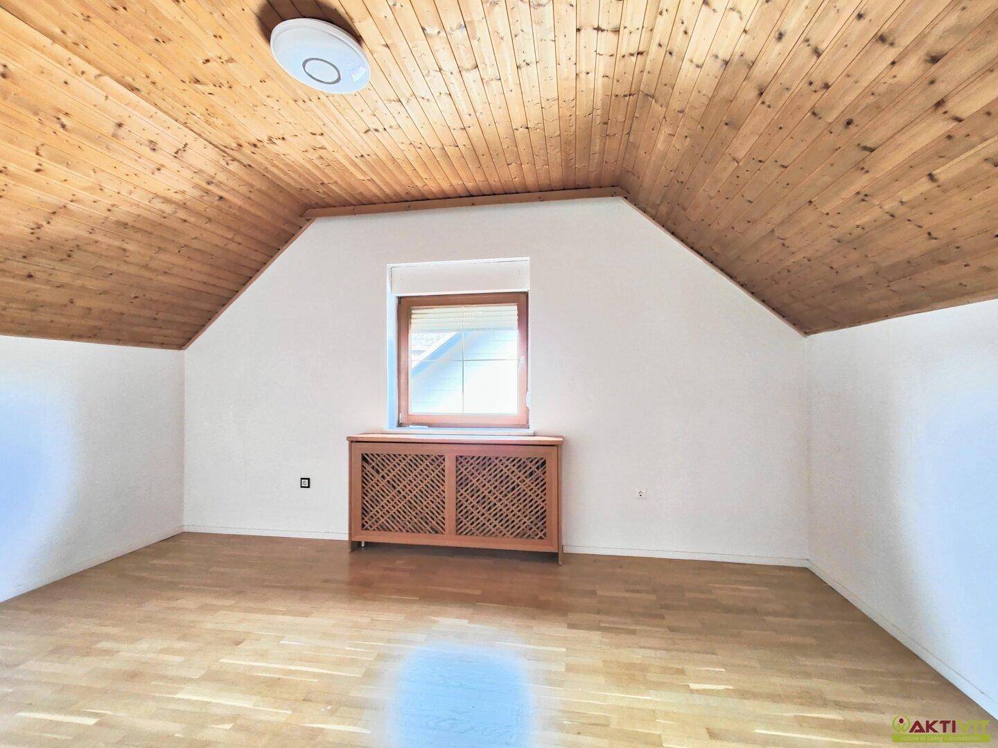 Geräumiges Dachzimmer mit Holzbalkendecke, Parkettboden und einem Fenster mit Heizungsverkleidung.
