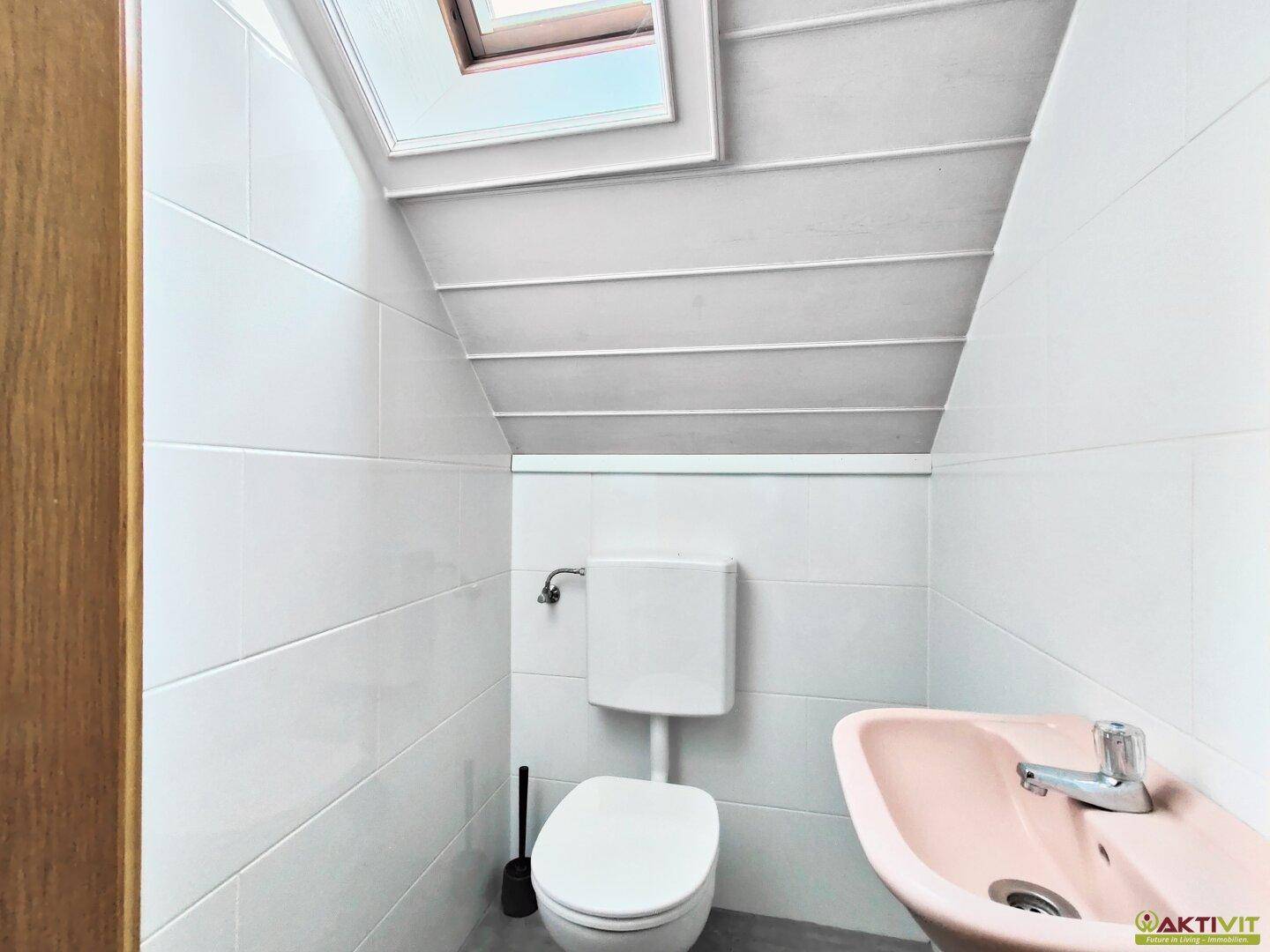 Separates WC mit Toilette, kleinem Waschbecken und Dachfenster in weiß gefliestem Raum.