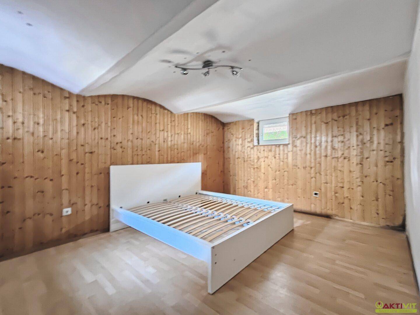 Schlafzimmer mit Holzwänden, Parkettboden und einem kleinen Fenster, bietet eine gemütliche Atmosphäre.