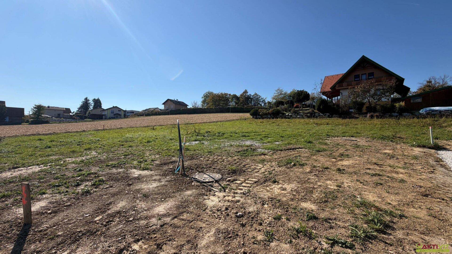 Blick auf ein sonniges Baugrundstück mit vorbereiteten Anschlüssen und einem Haus im Hintergrund.