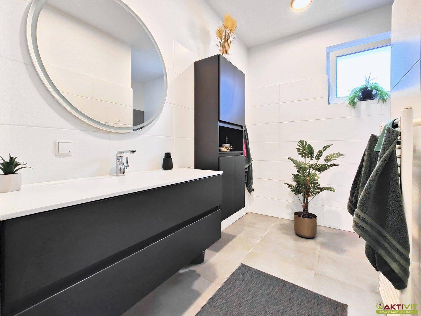 Modernes Badezimmer mit schwarzem Waschtisch, rundem Spiegel und Fenster für Tageslicht.