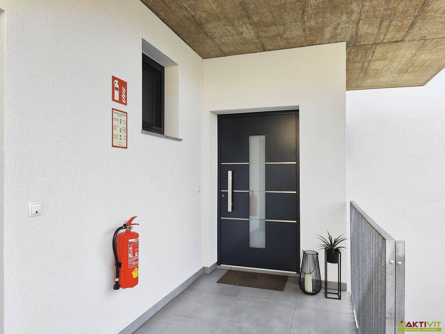 Eingangsbereich des Gebäudes mit moderner Tür, Feuerlöscher und kleinem Fenster.