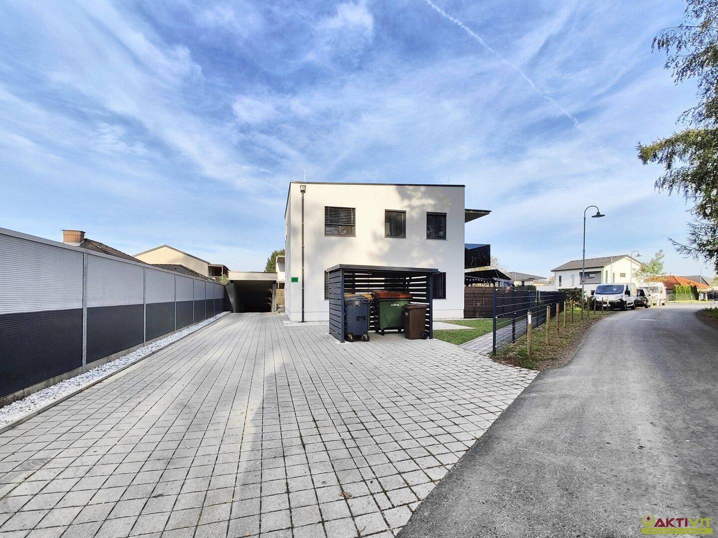 Seitenansicht des Gebäudes mit gepflasterter Zufahrt, Carport und Mülltonnenbereich.