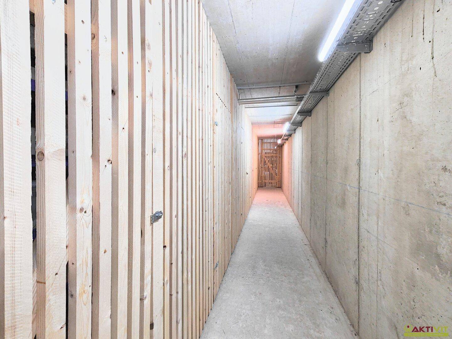 Langer Flur im Untergeschoss mit Betonwänden und Holzlatten-Abtrennungen für Lagerräume.