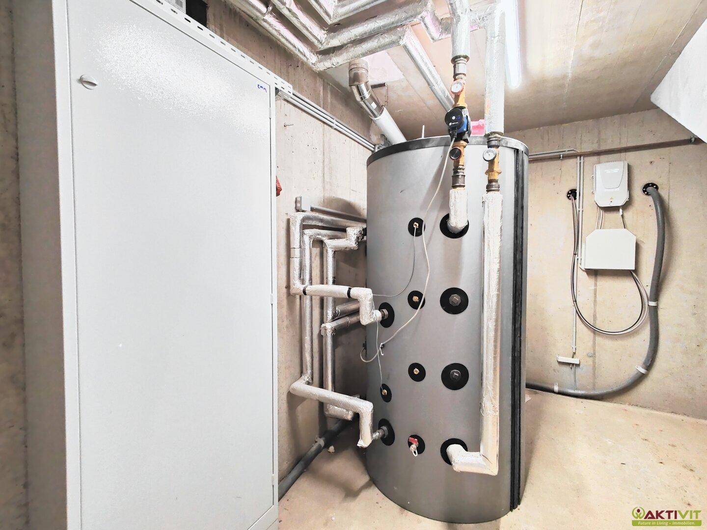 Technikraum im Keller mit großem Warmwasserspeicher, Rohrleitungen und einem Schaltkasten.