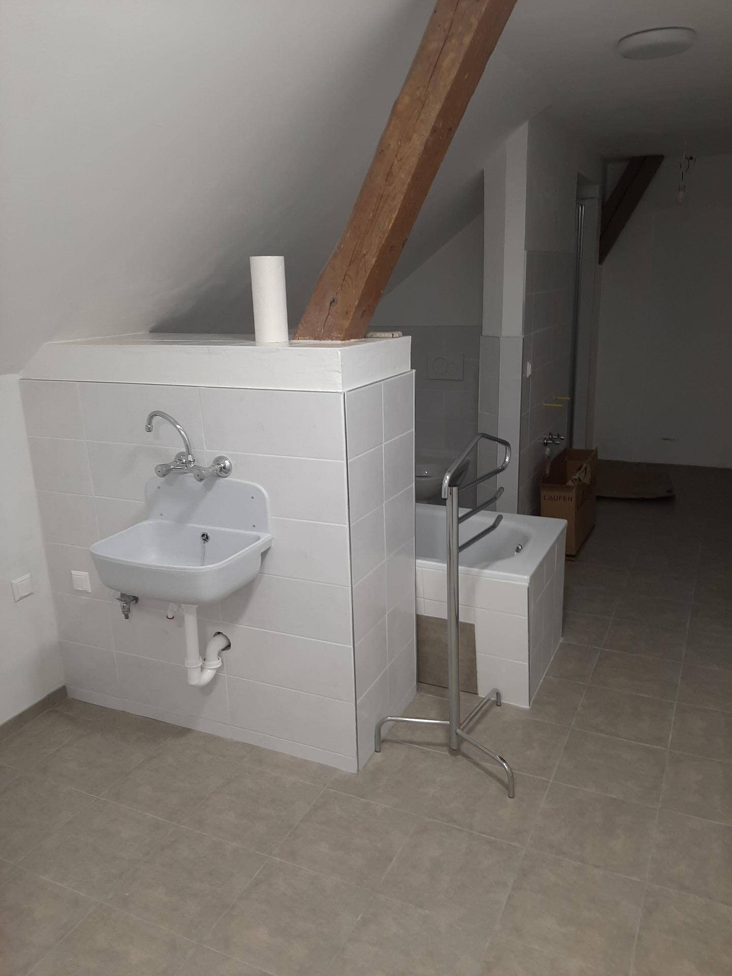 Badezimmerbereich mit kleinem Waschbecken, Badewanne und sichtbarem Holzbalken unter der Dachschräge.