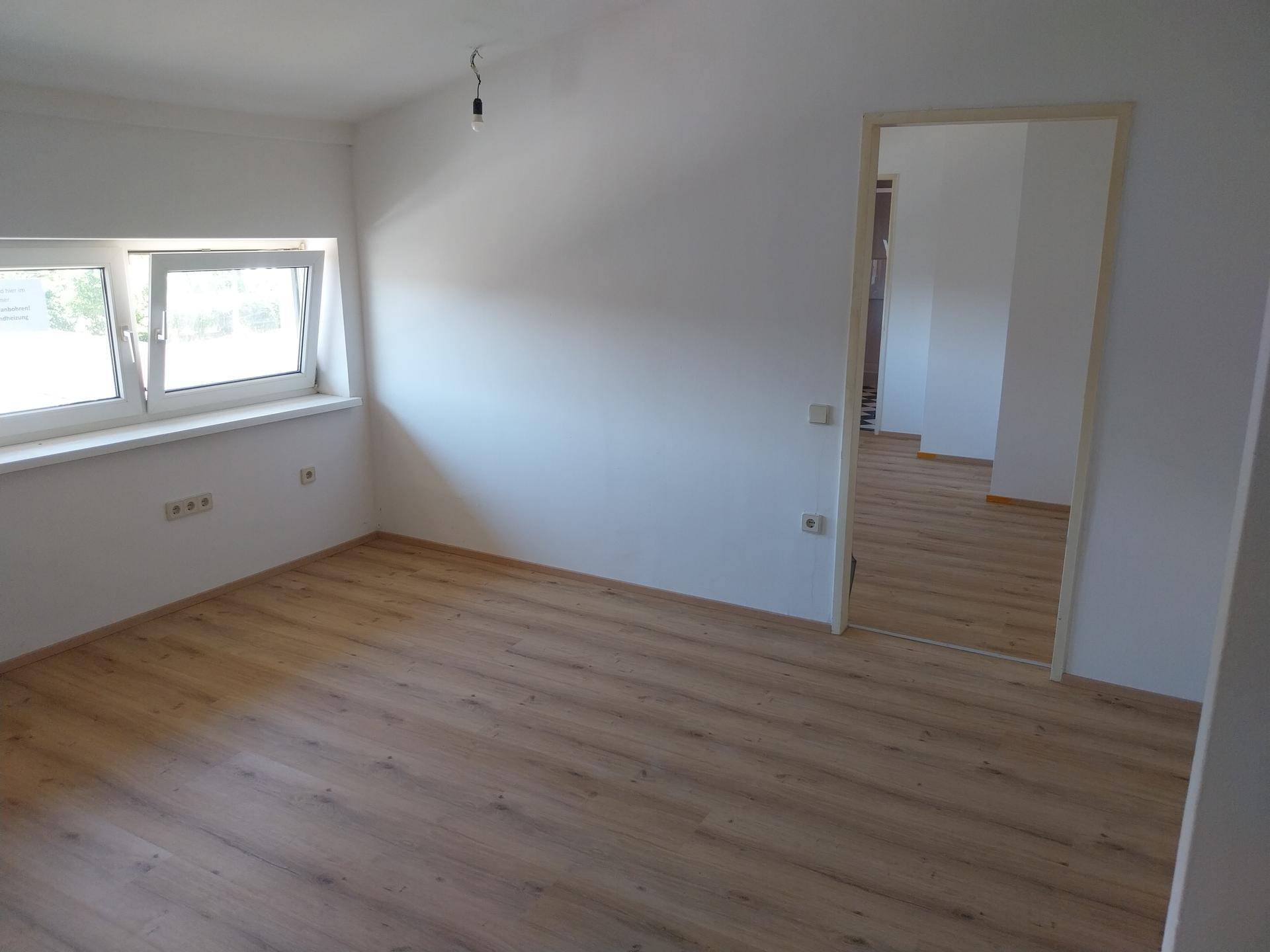 Leerer Raum mit hellem Laminatboden und weiß gestrichenen Wänden, mit Zugang zu einem weiteren Zimmer.