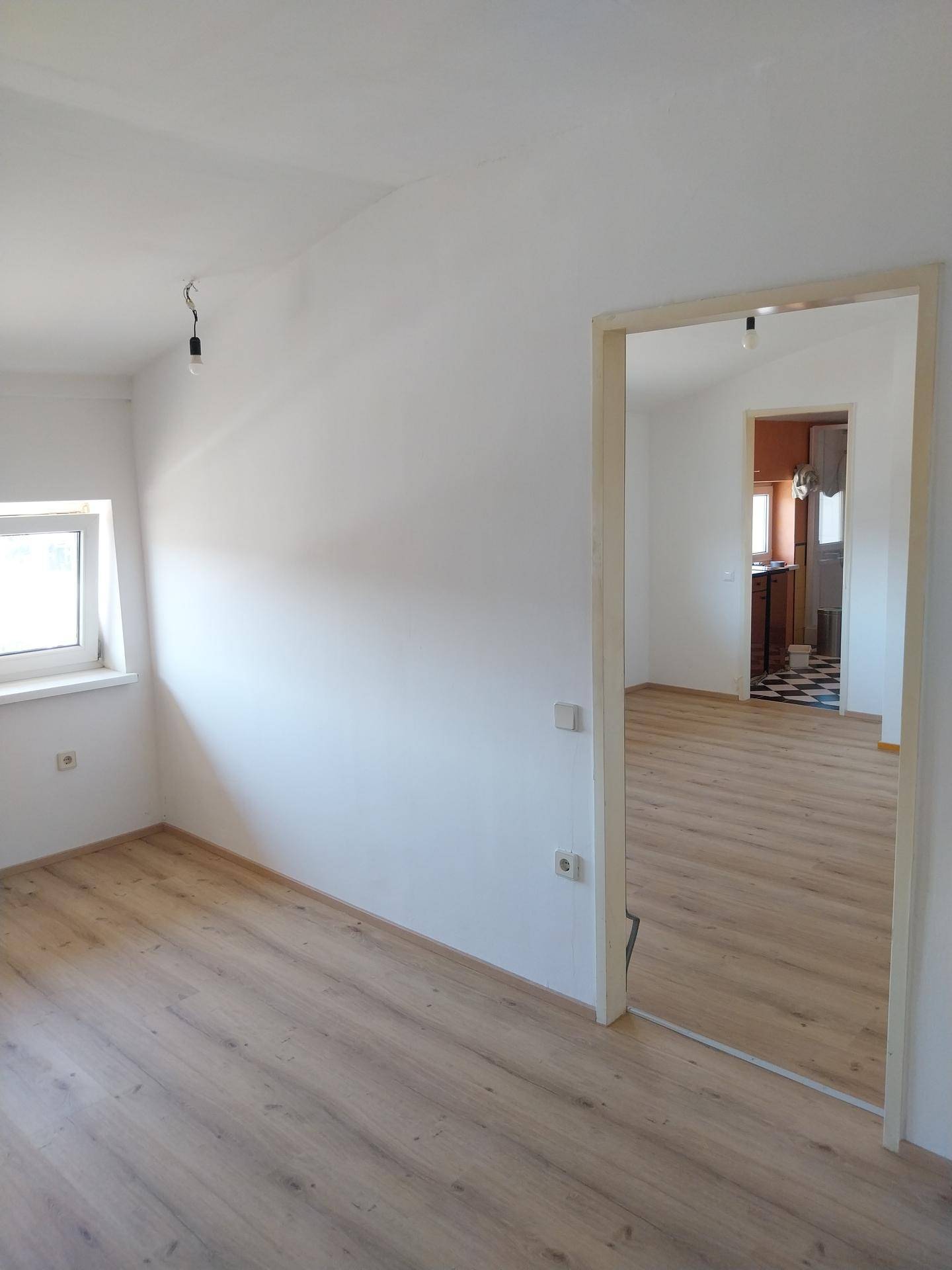 Heller Raum mit Laminatboden und weiß gestrichenen Wänden, mit Blick in ein angrenzendes Zimmer.