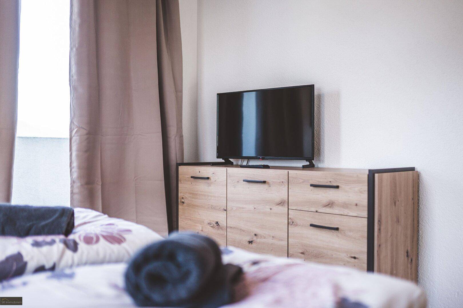 Detailansicht des Schlafzimmers mit TV auf einem Holzschrank und Vorhängen am Fenster.