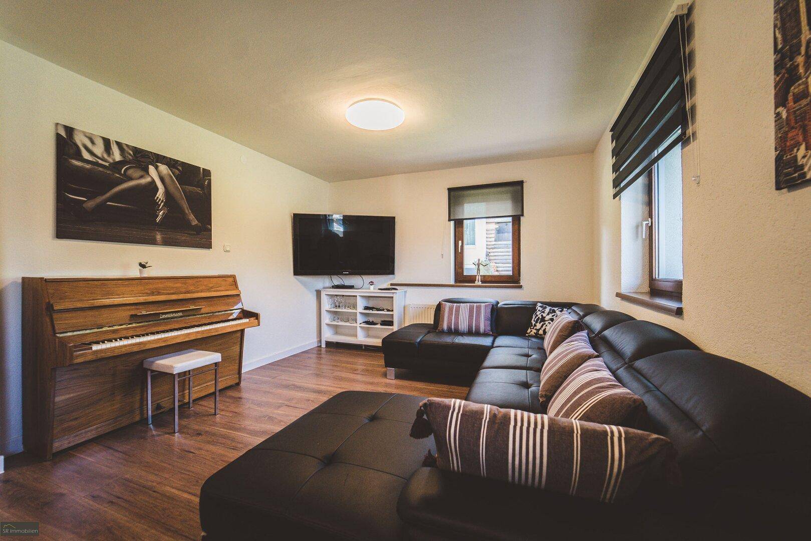 Geräumiges Wohnzimmer mit Ecksofa, Fernseher und Klavier, bietet viel Platz für Unterhaltung.