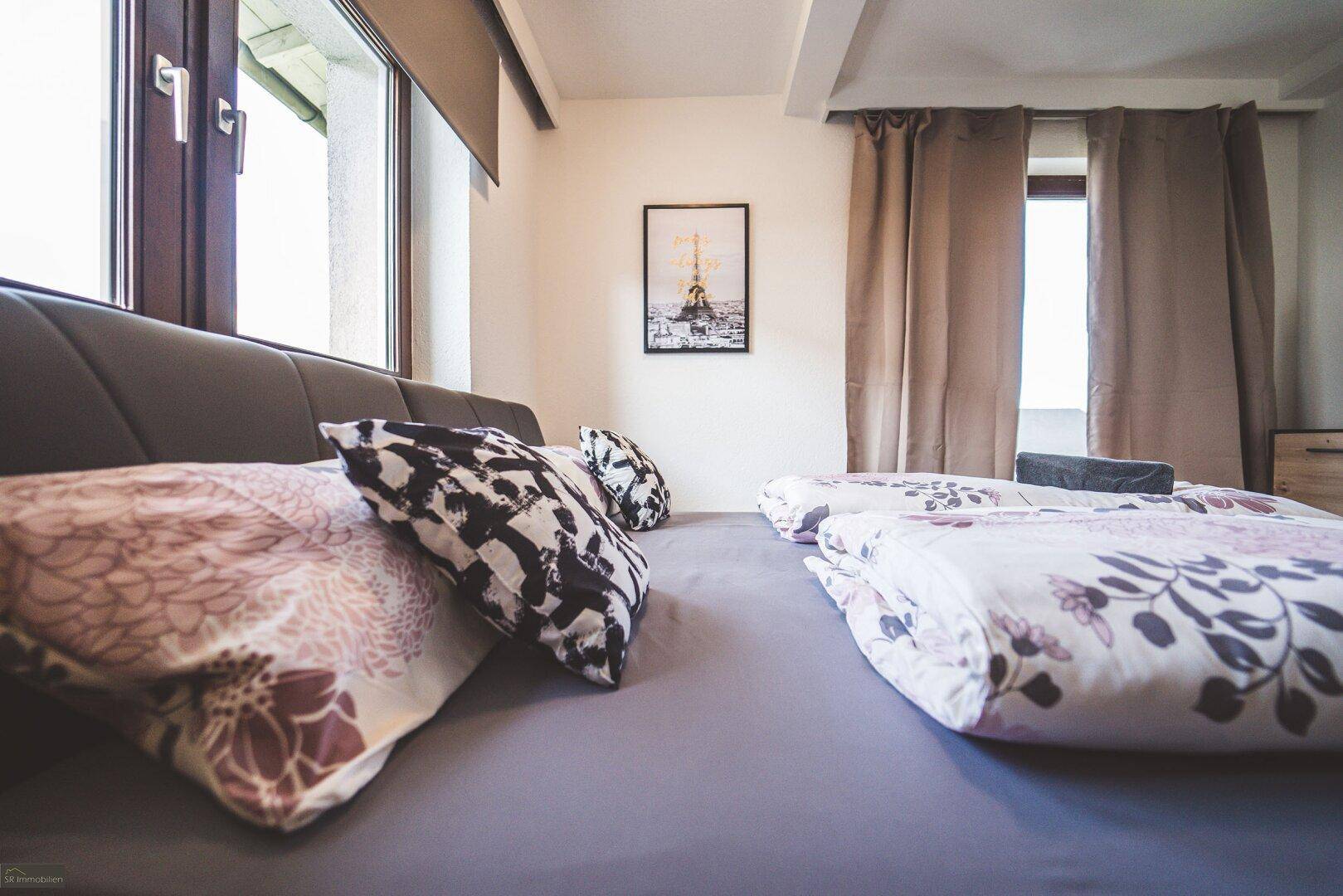 Nahaufnahme des Bettes im Schlafzimmer mit gemusterten Kissen und Vorhängen am Fenster.