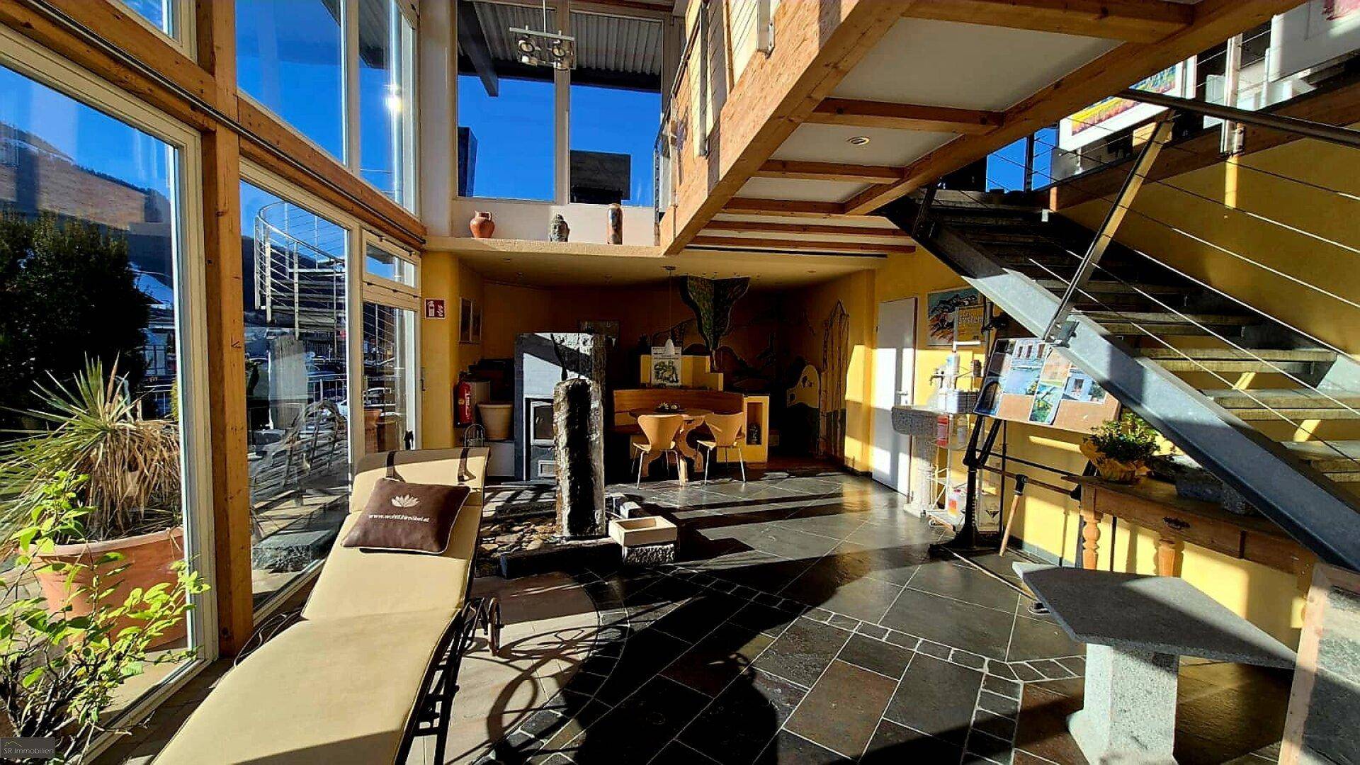 Heller Showroom mit bequemer Liege, Pflanzen und einer Holztreppe, die zu den oberen Etagen führt. Große Fenster bieten viel Tageslicht.