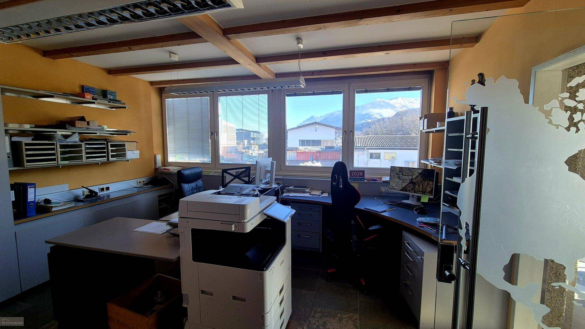 Funktionales Büro mit Schreibtisch, Computer, Drucker und Regalen, bietet einen klaren Bergblick durch das Fenster.