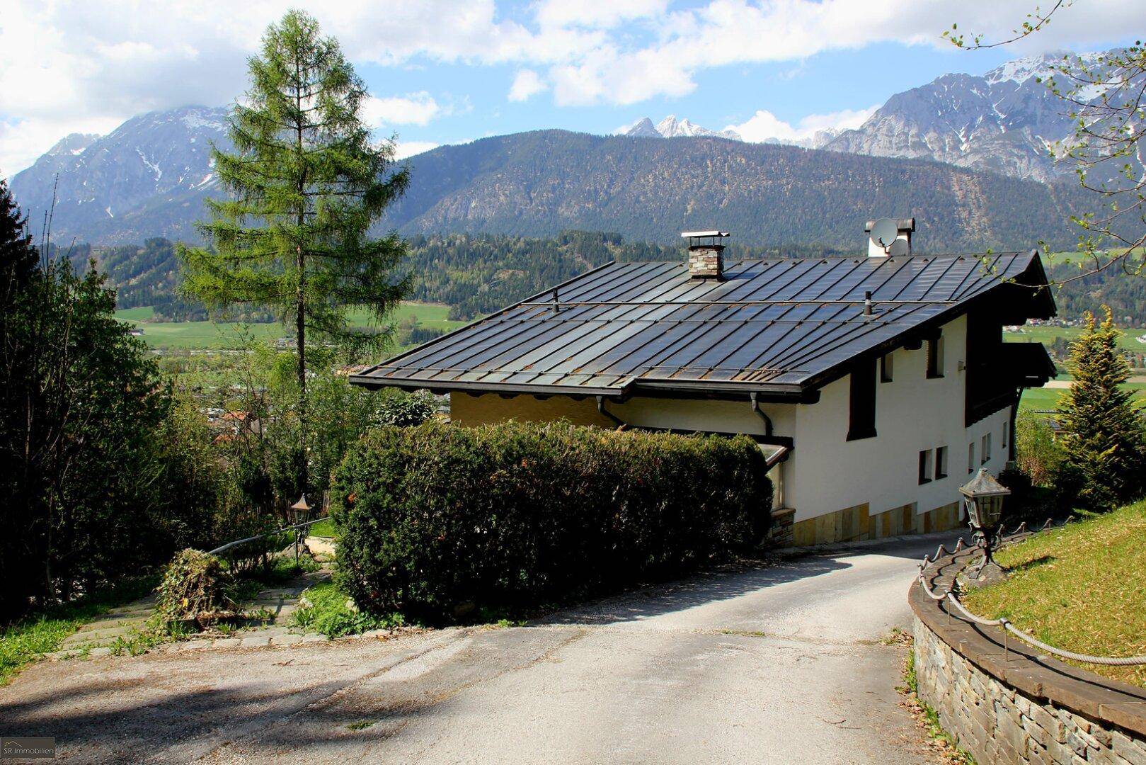 Haus mit Photovoltaikanlage auf dem Dach und Zufahrt, umgeben von grüner Natur und Bergen.