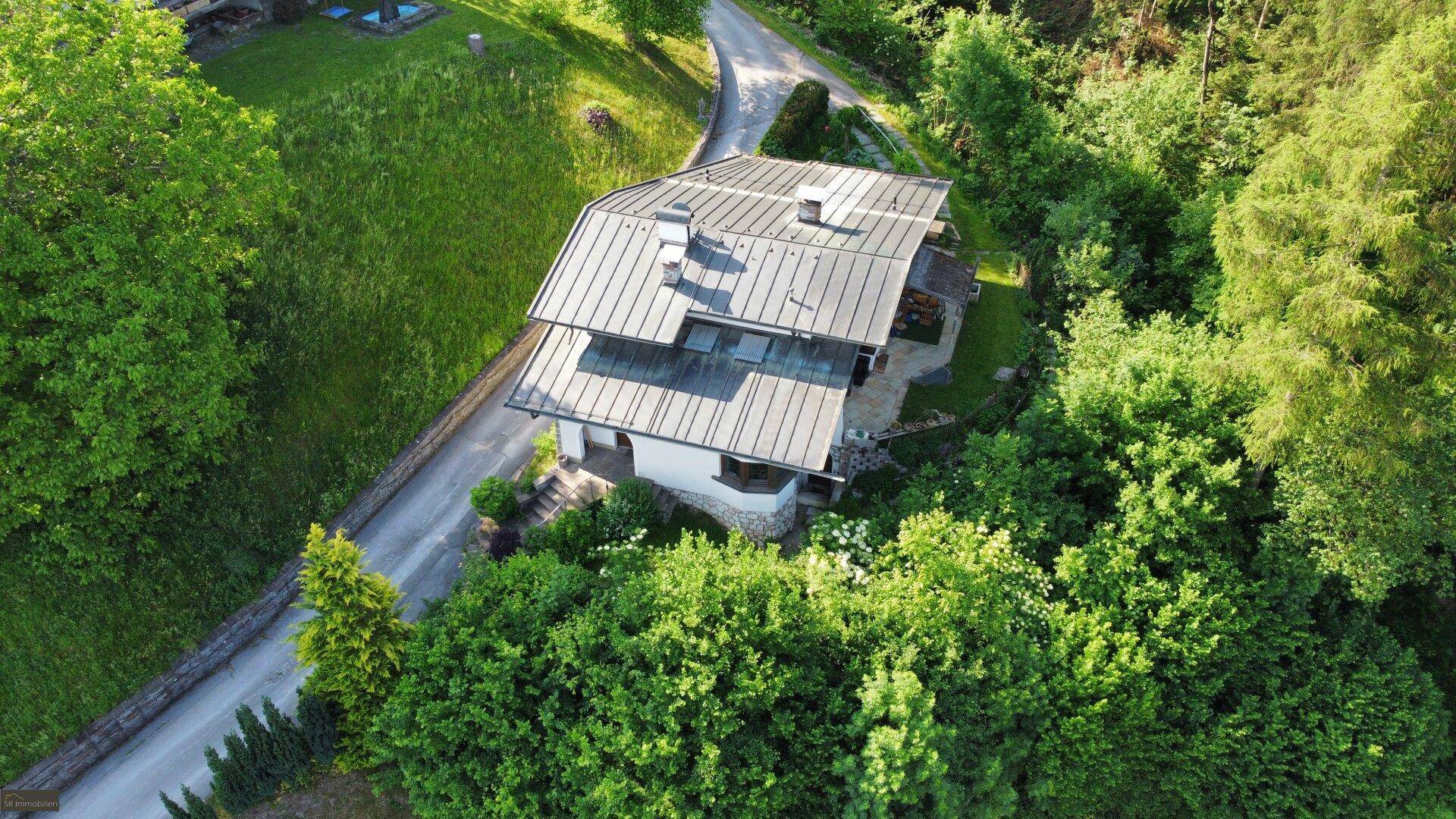 Idyllische Luftaufnahme eines Hauses im Grünen, umgeben von Bäumen und einer geschwungenen Zufahrt.