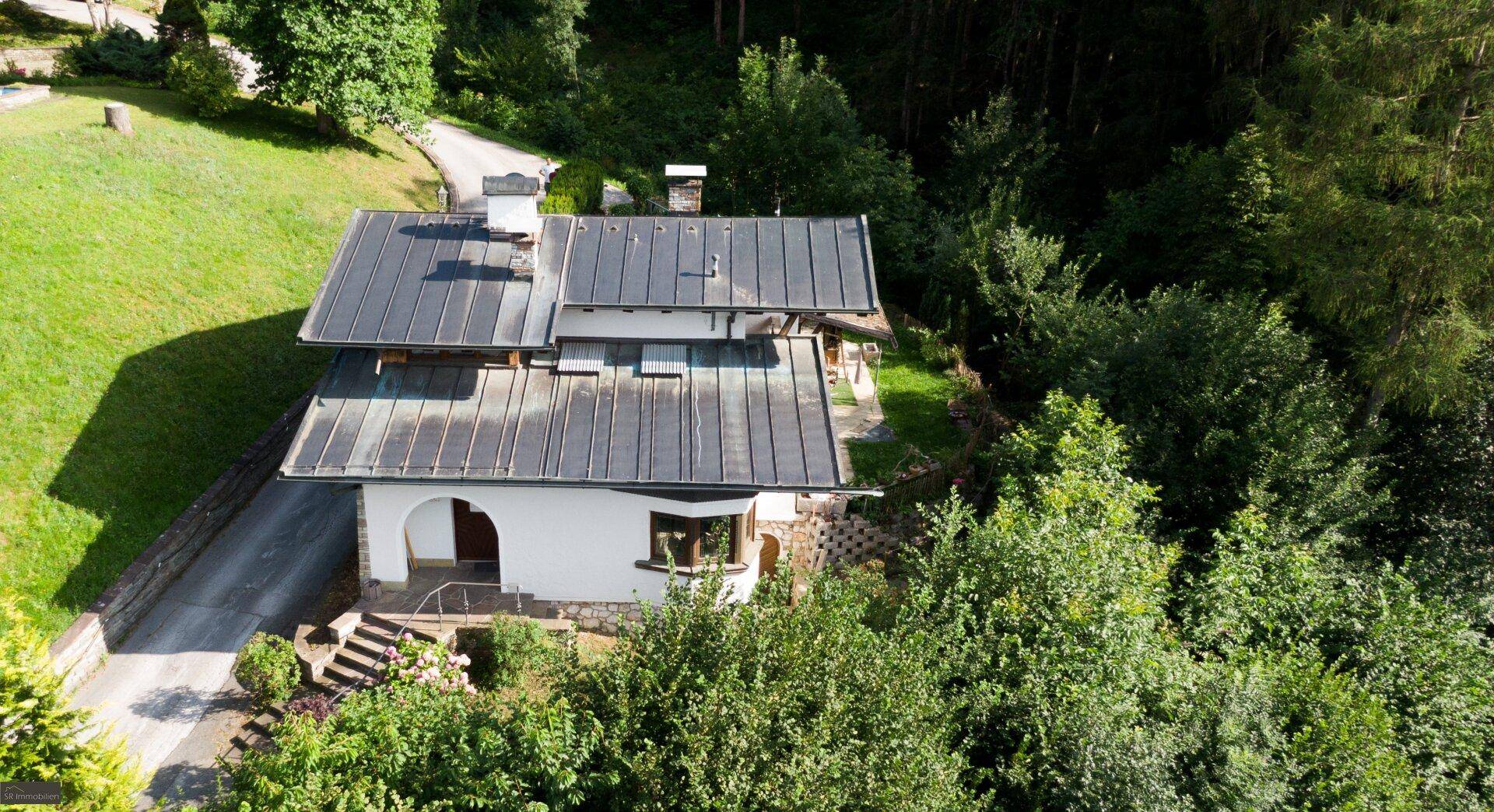 Vogelperspektive eines charmanten Hauses mit dunklem Dach und weißer Fassade, eingebettet in eine grüne, bewaldete Umgebung.