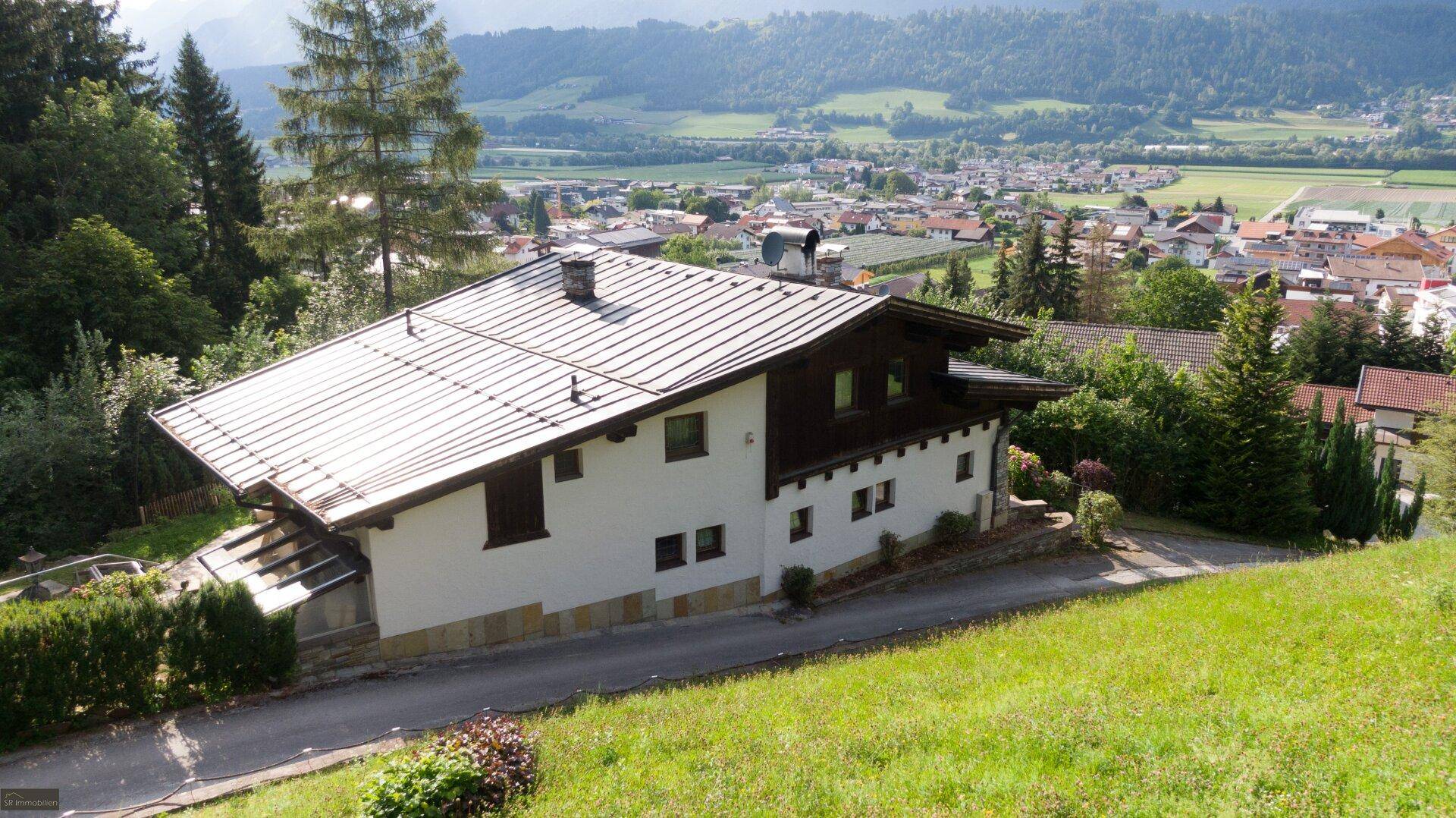 Seitenansicht des Hauses mit weißer Fassade und dunklem Dach, umgeben von grünen Hügeln und einem Talblick.