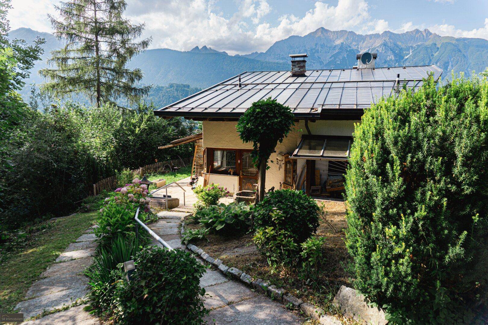 Gepflegter Gartenbereich mit Steinweg, umgeben von Pflanzen und Büschen, mit Bergblick im Hintergrund.
