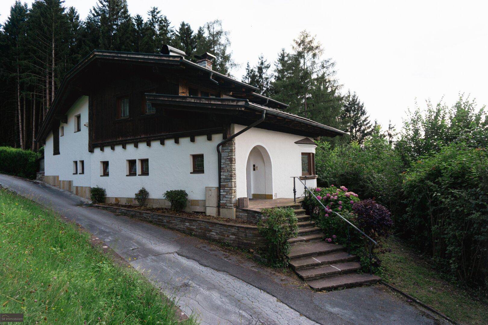 Eingangsbereich des Hauses mit Steintreppe und einem gewölbten Eingang, umgeben von üppigem Grün und Bäumen.