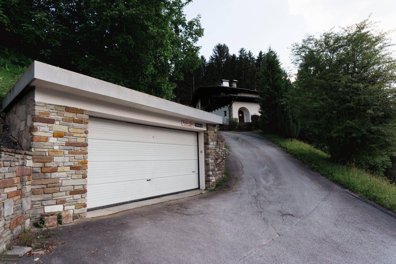 Separate Garage mit weißem Rolltor und Steinverkleidung, mit einer Auffahrt zum Haupthaus im Hintergrund.