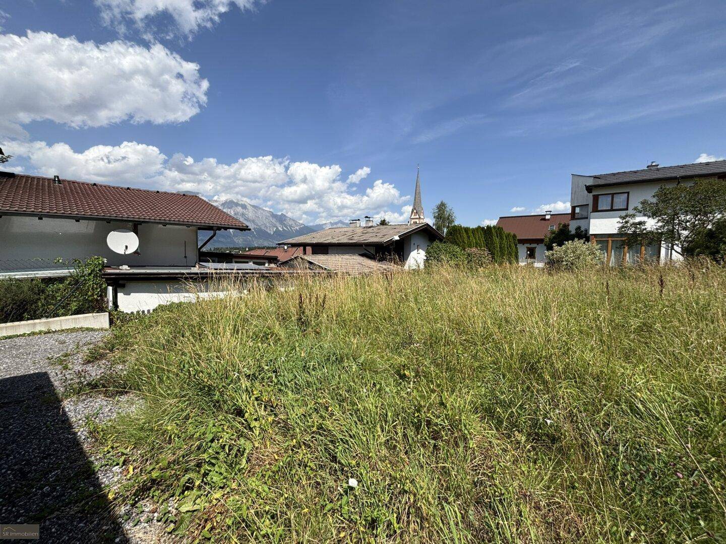 Blick über ein grünes Grundstück mit Schotterweg, umgeben von Häusern und einer beeindruckenden Bergkulisse.
