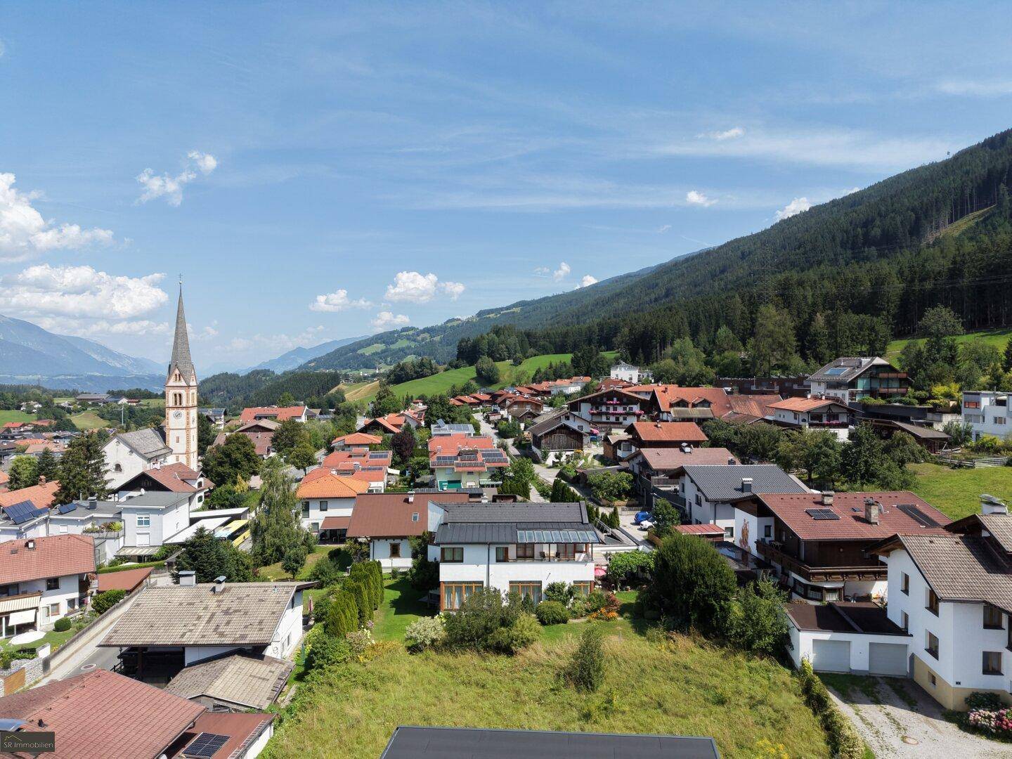 Panoramablick auf ein malerisches Dorf mit Kirchturm, umgeben von grünen Hügeln und Bergen.