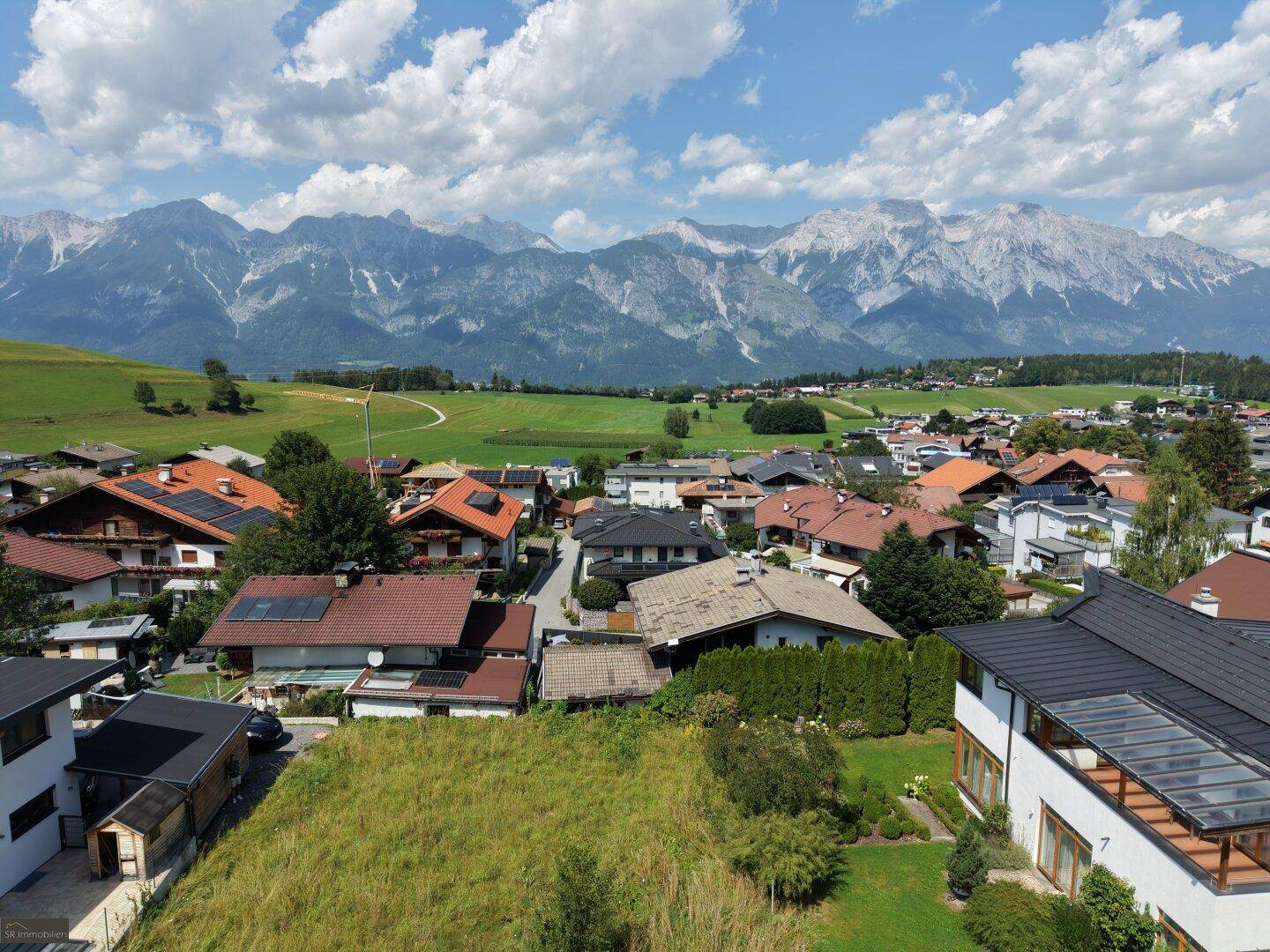 Weitläufiger Blick über das Dorf mit dem unbebauten Grundstück im Vordergrund und majestätischen Bergen im Hintergrund.