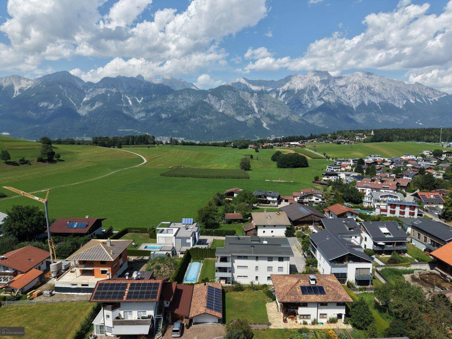 Breiter Panoramablick auf die ländliche Umgebung mit Wohnhäusern, Feldern und einer beeindruckenden Bergkulisse.