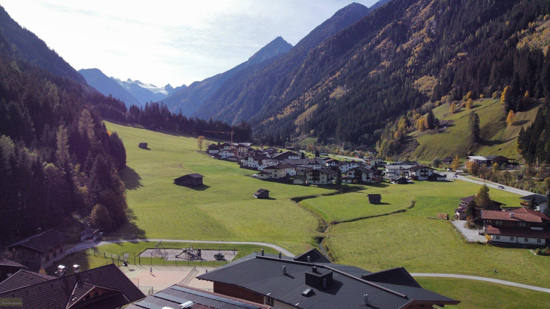 Panoramablick auf ein idyllisches Alpendorf in einem weiten Tal, umgeben von grünen Wiesen und hohen Bergen.