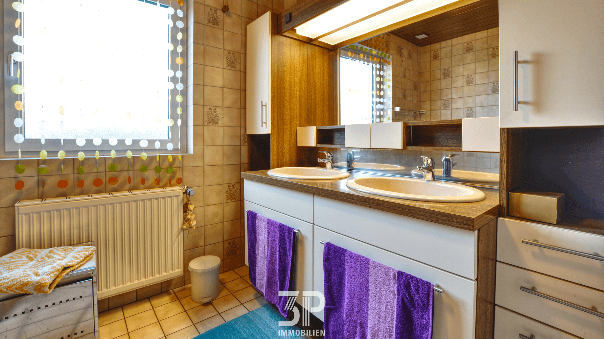 Gefliestes Badezimmer mit Doppelwaschbecken, Spiegelschrank und Fenster.