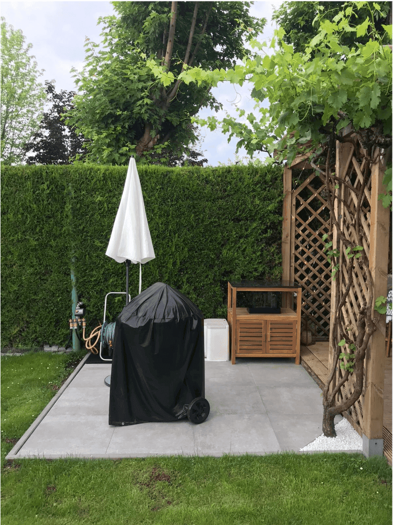 Gartenbereich mit Grillplatz, Sonnenschirm und Holzpergola, umgeben von einer hohen Hecke.