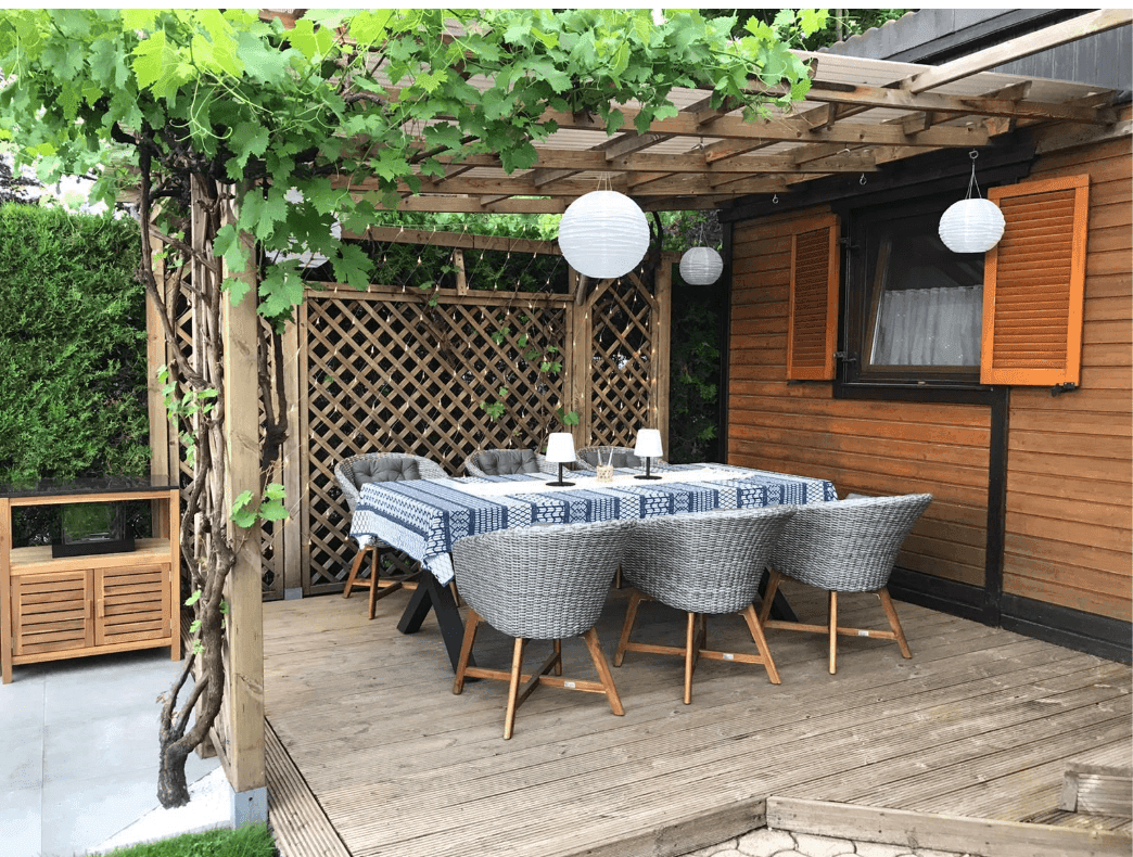 Sonnige Holzterrasse mit Esstisch und Korbstühlen, umgeben von grünen Pflanzen und Hecken.