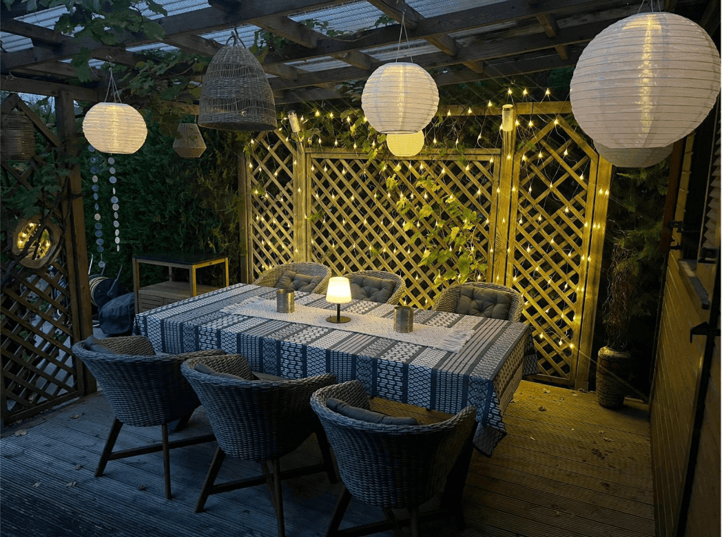 Beleuchtete Holzterrasse mit Esstisch und Korbstühlen, ideal für Abendessen im Freien.