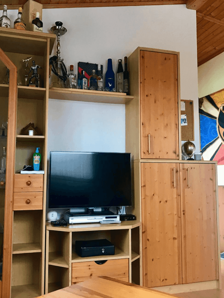 Wohnzimmer mit Holzregal, Fernseher und dekorativen Elementen unter einer Holzbalkendecke.