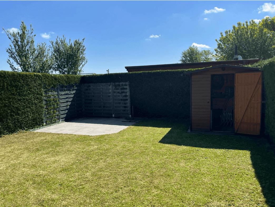 Sonniger Garten mit Rasenfläche, Gartenhaus und gepflastertem Bereich, umgeben von Hecken.