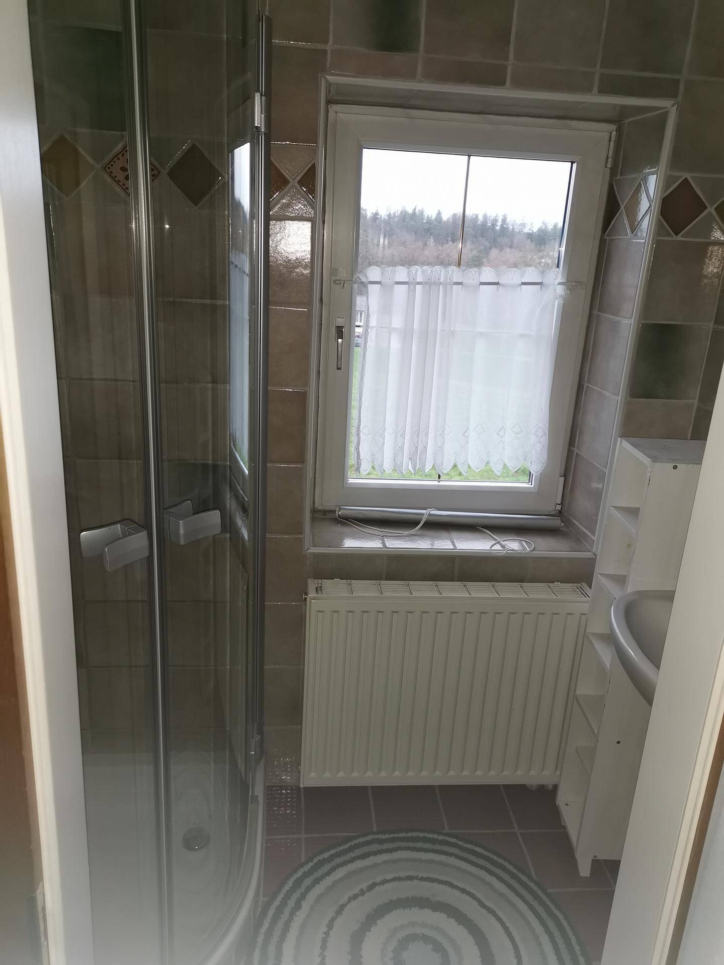 Kleines Badezimmer mit Duschkabine, Waschbecken, Heizkörper und Fenster.