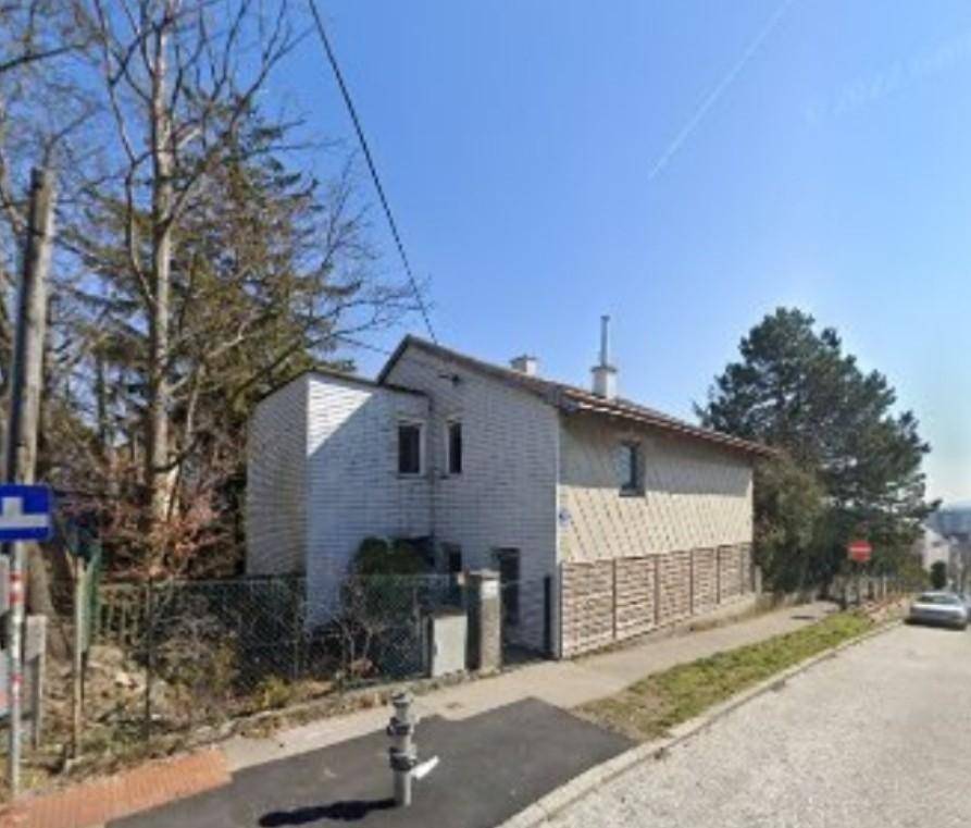 Hausfassade von der Straße aus gesehen mit angrenzendem Gehweg und Bäumen im Hintergrund.