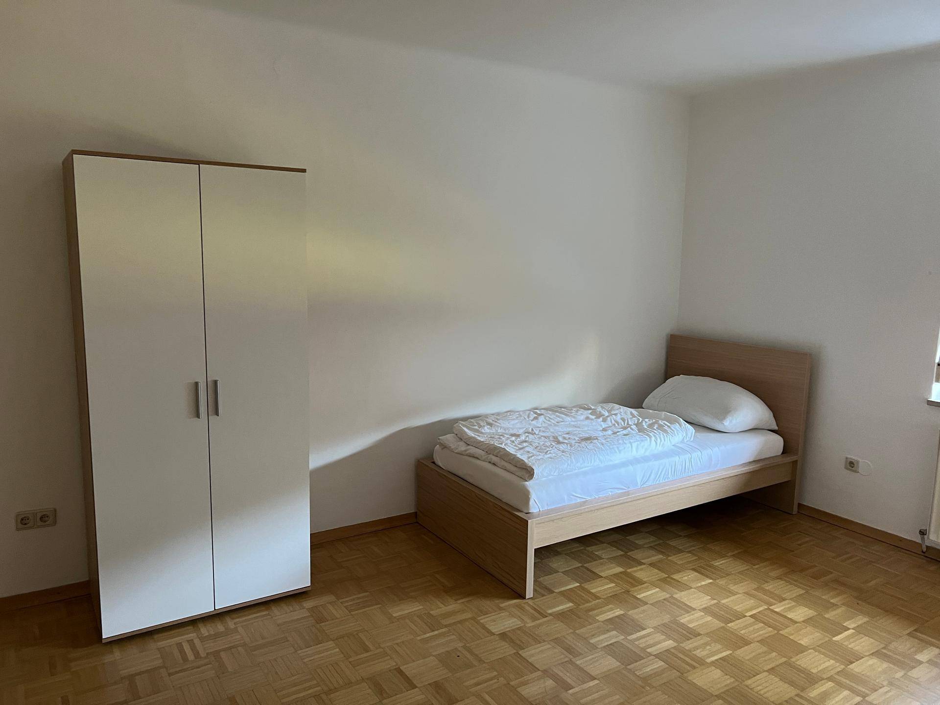 Einfaches Schlafzimmer mit Einzelbett, weißem Kleiderschrank und Parkettboden, gut beleuchtet.