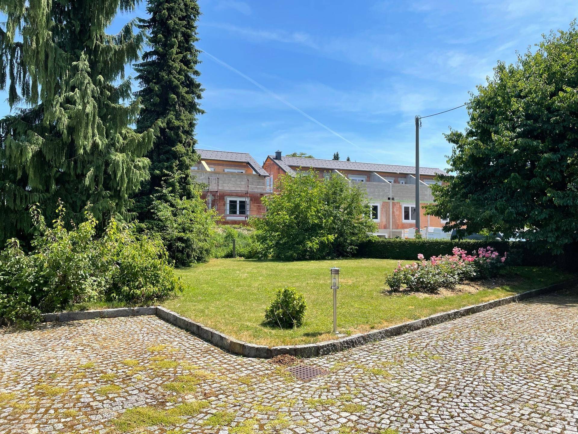 Gepflegter Gartenbereich mit Rasenfläche und Sträuchern, umgeben von Bäumen und Pflastersteinen.