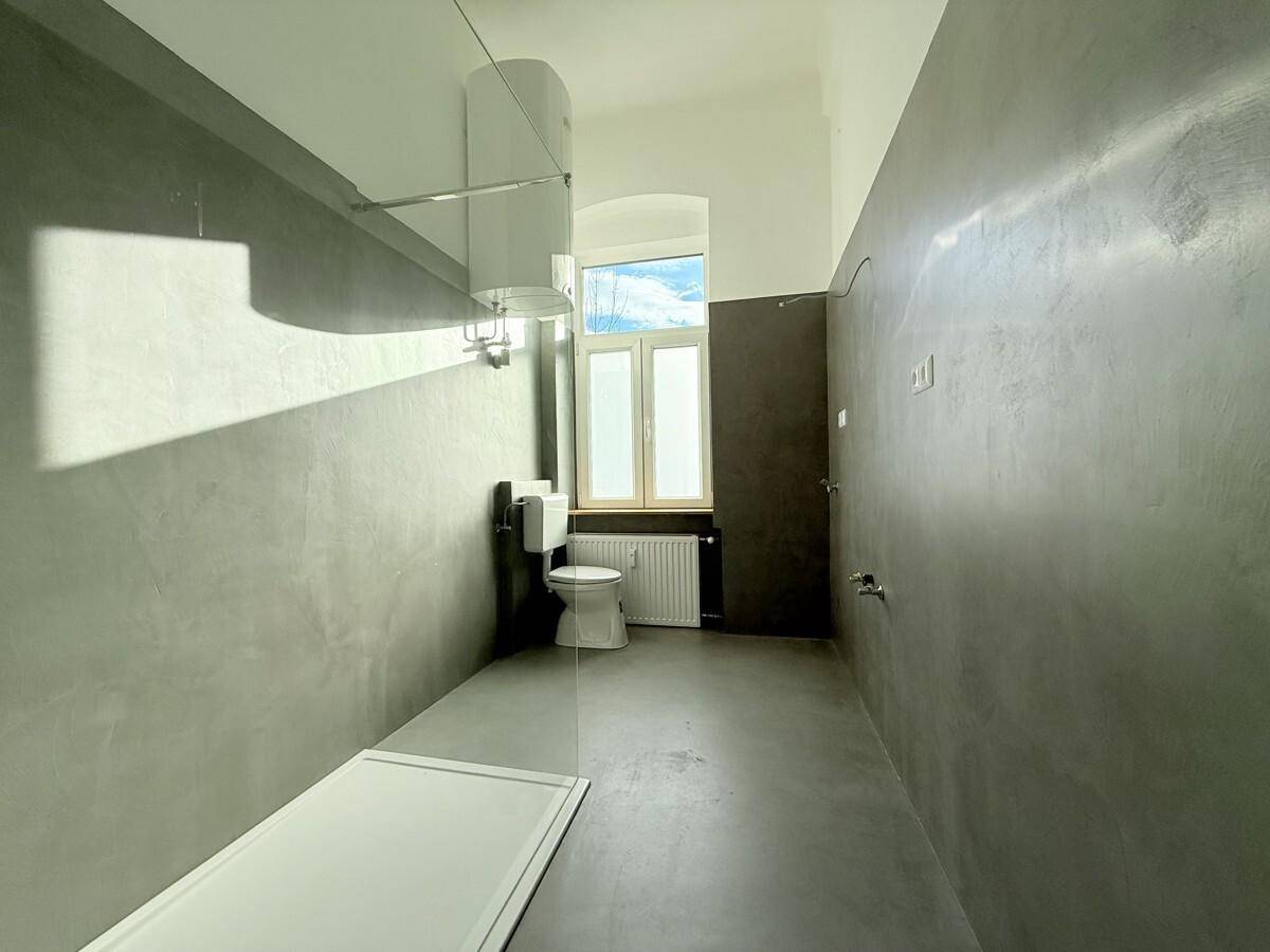 Modernes Badezimmer mit grauen Wänden, Toilette und Duschbereich, gut beleuchtet durch ein Fenster.