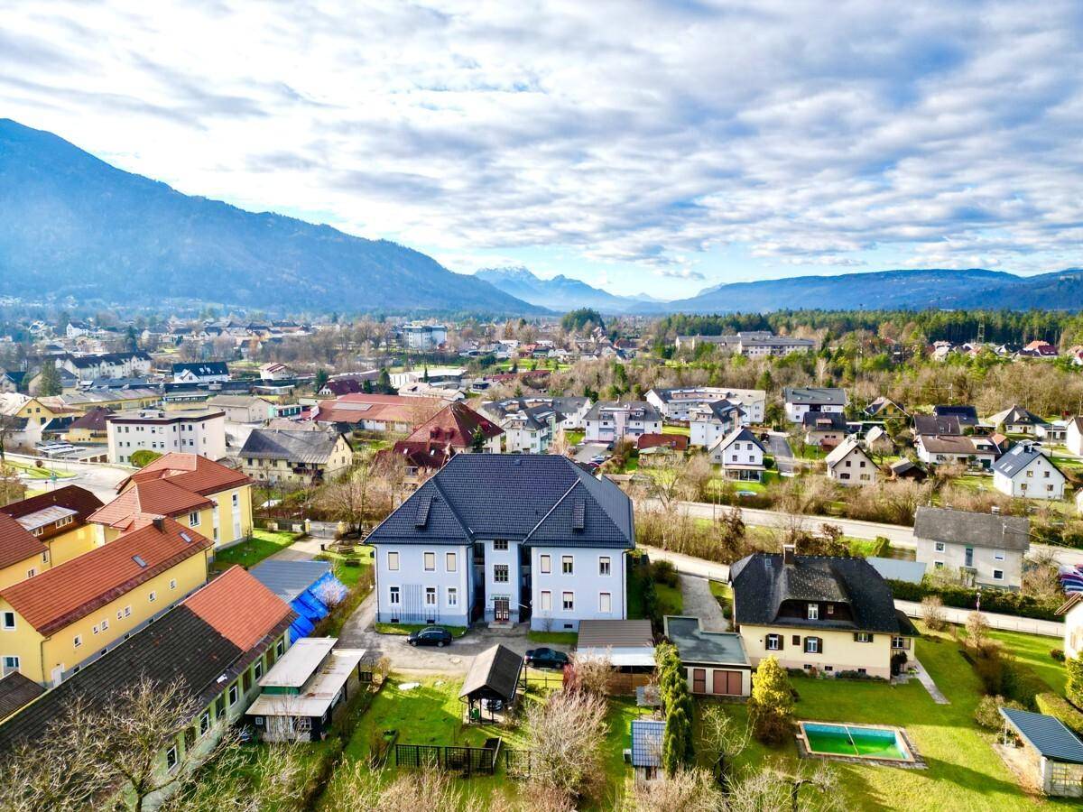 Luftaufnahme eines Wohngebäudes in einer ländlichen Umgebung mit Bergen im Hintergrund unter bewölktem Himmel.