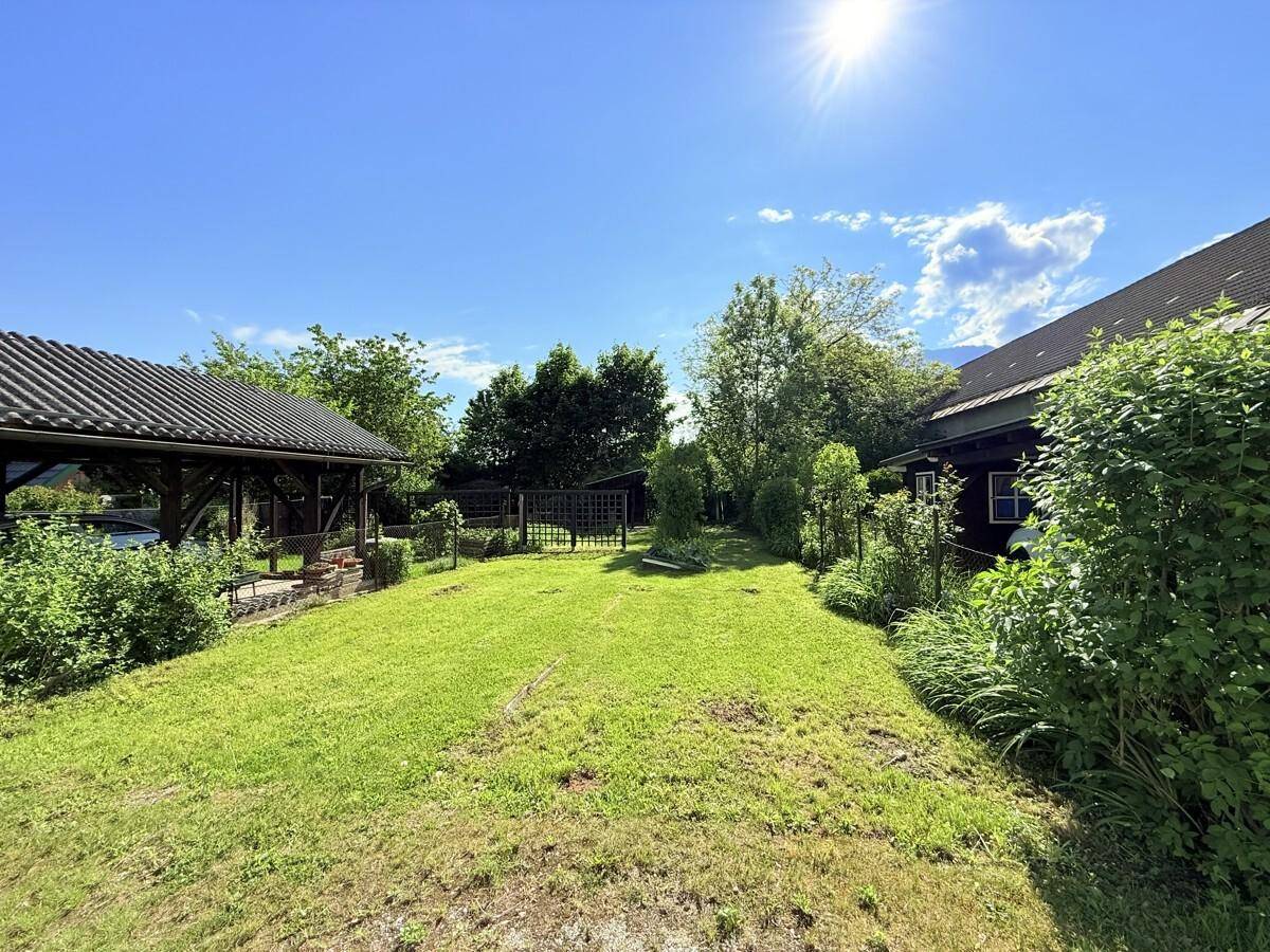 Sonniger Gartenbereich mit Rasenfläche, Bäumen und einer überdachten Terrasse im Hintergrund.