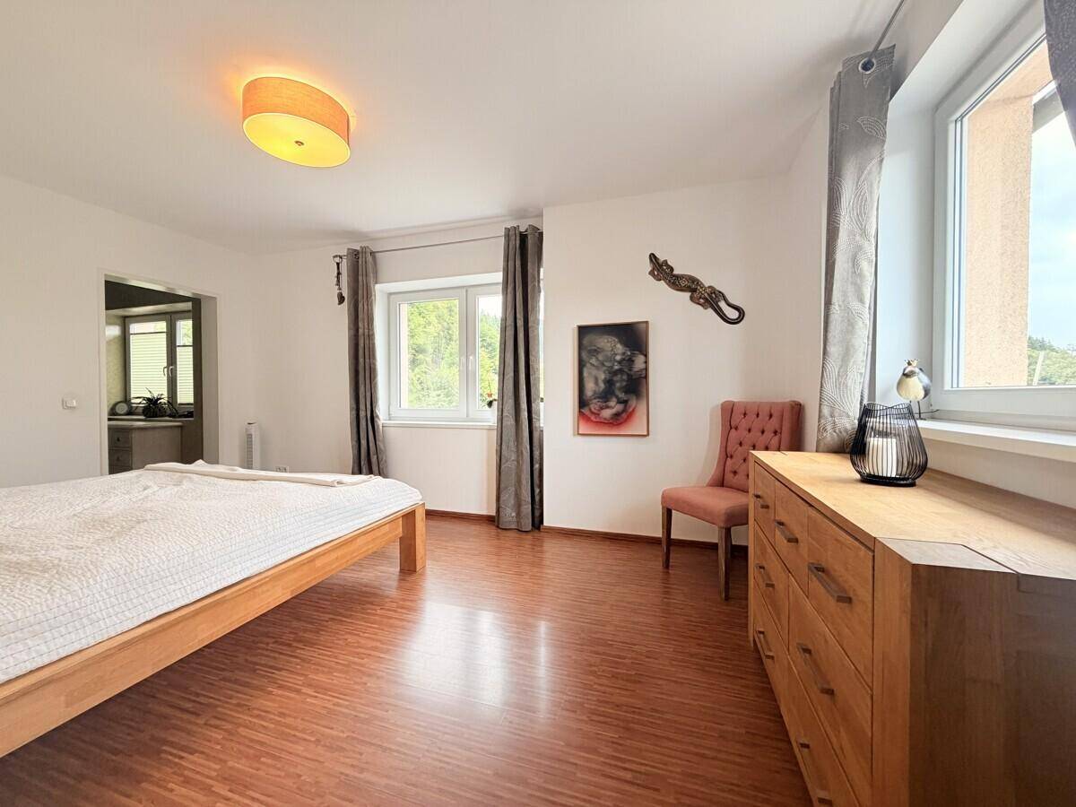 Helles Schlafzimmer mit Holzbett, Kommode und Fenstern mit Blick ins Grüne.