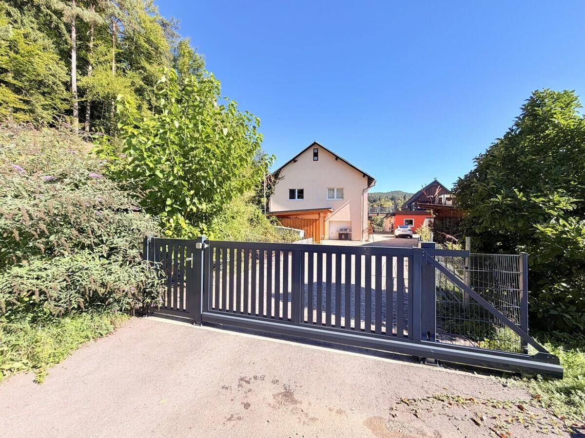 Modernes Einfahrtstor aus Metall vor einem Wohnhaus, umgeben von üppigem Grün.