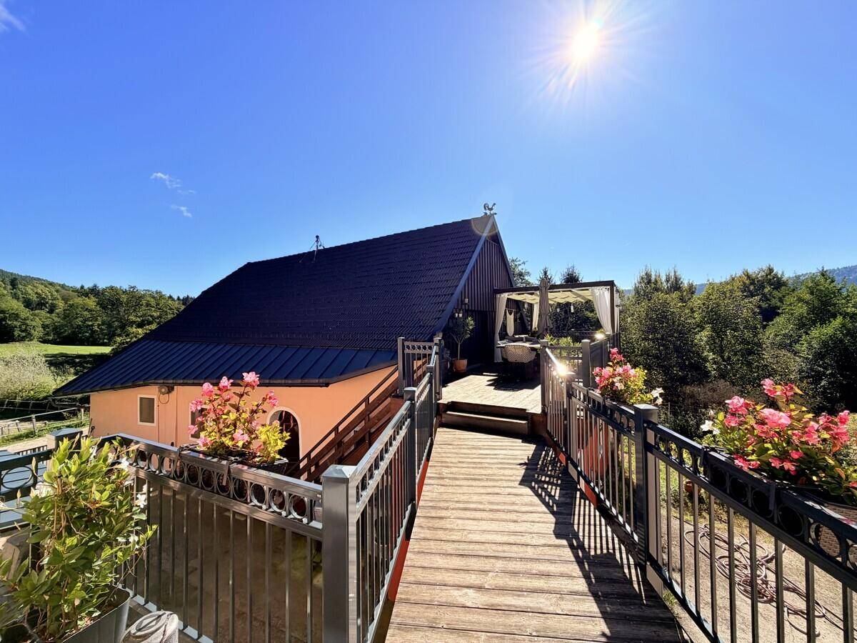Hausfassade mit dunklem Dach und einem Holzsteg, der zu einer erhöhten Terrasse mit weißem Sonnenschutz führt.