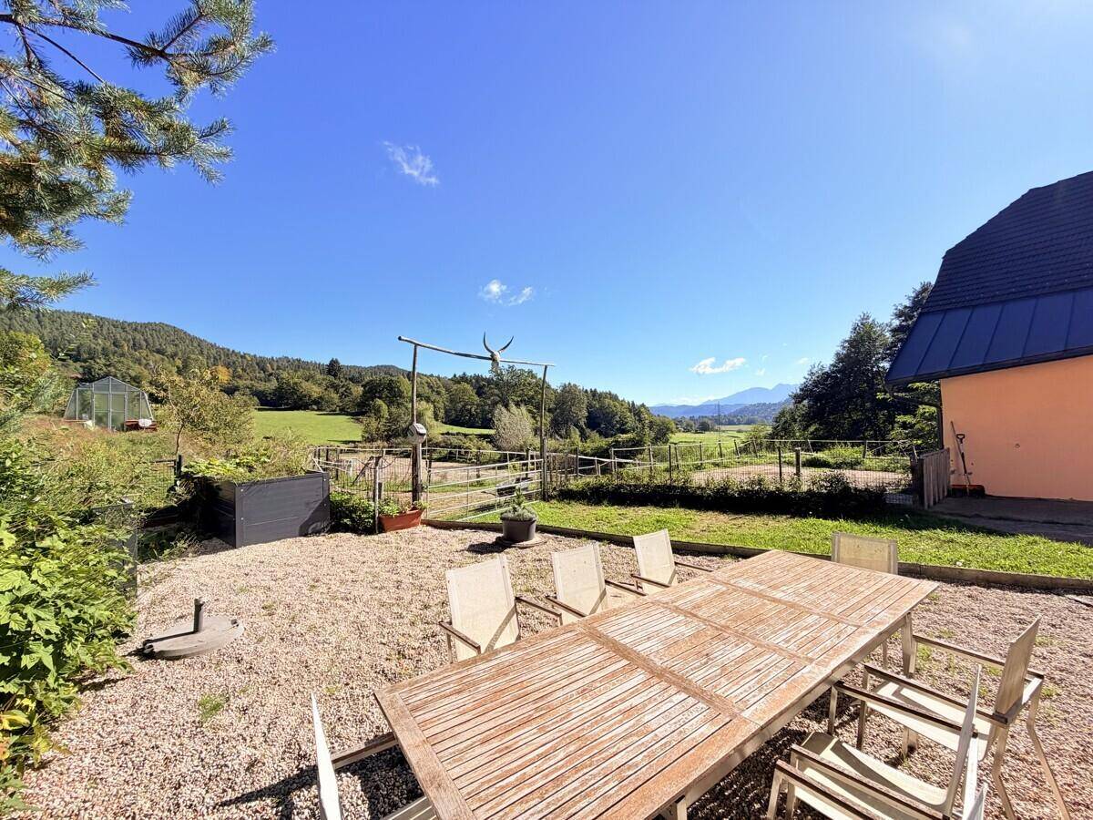 Gemütliche Terrasse mit Holztisch und Stühlen auf Kies, umgeben von ländlicher Natur und Bergblick.